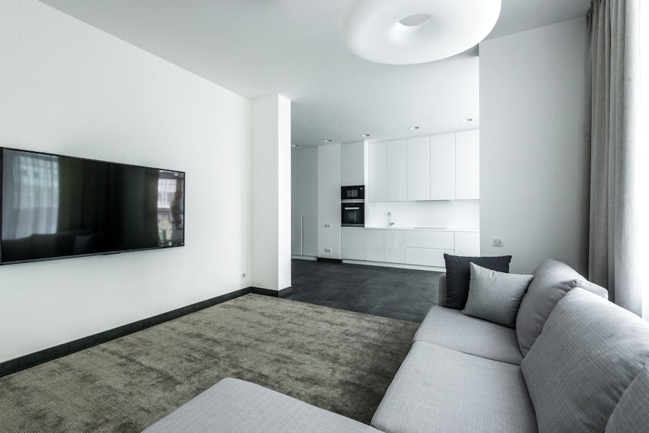 A modern North London flat living room featuring a light grey sofa with matching cushions placed on a textured beige carpet. The wall-mounted flat-screen TV is on a white wall opposite the sofa, with 