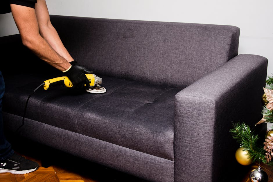 A person wearing black gloves is using a handheld upholstery cleaning device connected to a power cord to deep clean a dark grey fabric sofa in a residential setting. The sofa has a modern design with
